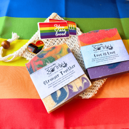 Alternate view flatlay photo showing items included in the Pride soap sampler - two half bars of Pride Month soap, a sisal fiber soap saver bag, two stickers, and a lapel pin