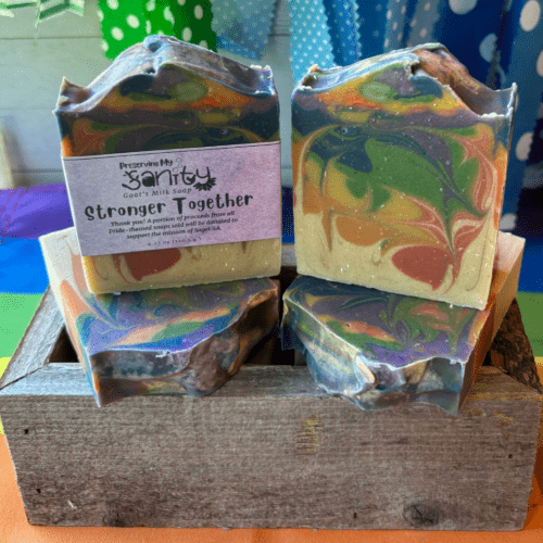 Photo of colorful rainbow drop swirl goat's milk soap to celebrate Pride Month. The name of the soap is "Stronger Together" and the photo features four bars displayed at different angles with a colorful backdrop.