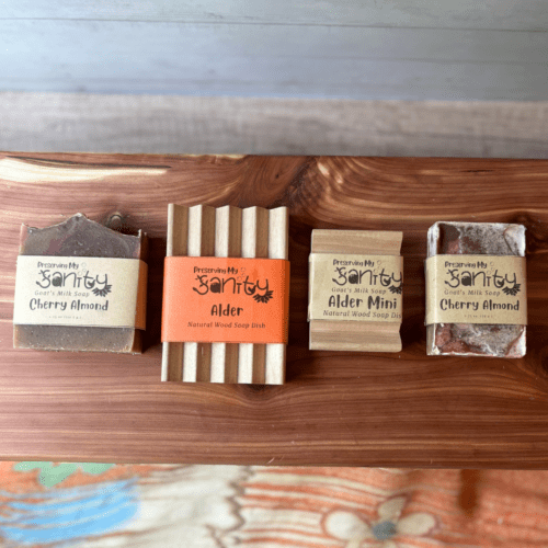 Flatlay photo showing a regular size bar of cherry almond goat soap, a regular size alder natural wood soap dish, and a smaller bar of Cherry Almond goat milk soap with an alder mini soap dish. Purpose of photo is to show scale/size of the different soap dishes.