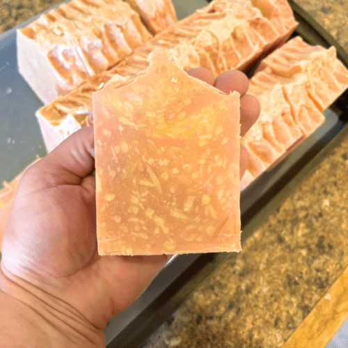 Process photo showing maker's hand holding a bar of freshly cut strawberry lemonade goat milk soap, with other freshly cut bars in background