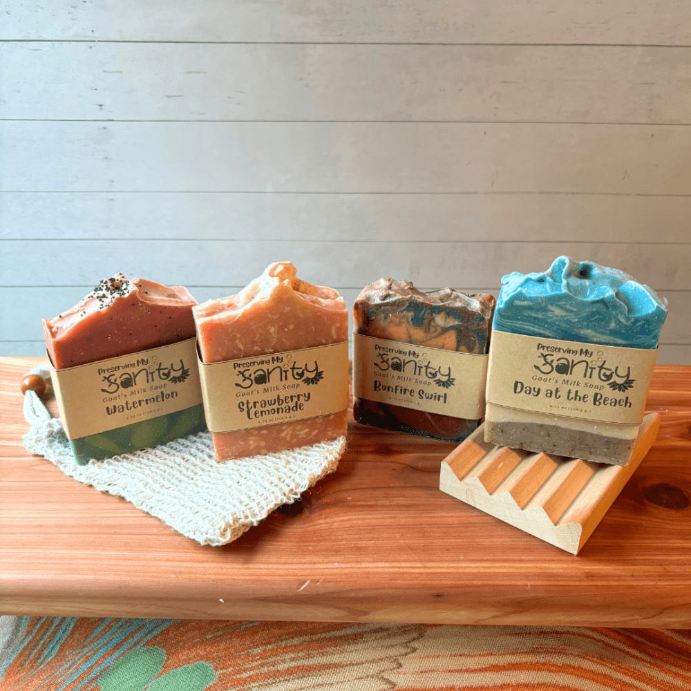 Side view flatlay photo showing four full size bars of summer scents of goat milk soap, with a soap saver bag and natural wood soap dish - all of these items are sold in the summer soap bundle. The soaps in this photo have labels on, so the middle part of the design is hidden.