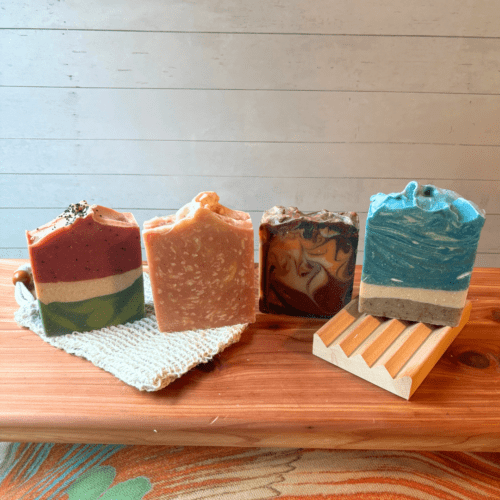 Alternate side view flatlay photo showing four full size bars of summer scents of goat milk soap, with a soap saver bag and natural wood soap dish - all of these items are sold in the summer soap bundle. The soaps in this photo don't have labels so customers can see full soap design.