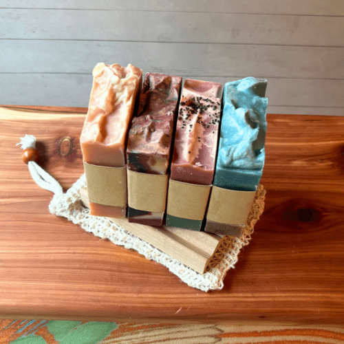 Overhead alternate view of the four full size soaps that are in the summer soap bundle - Day at the Beach, Strawberry Lemonade, Watermelon, and Bonfire. Pictured on top of a sisal soap saver bag and natural wood soap dish, which are also included in the bundle.