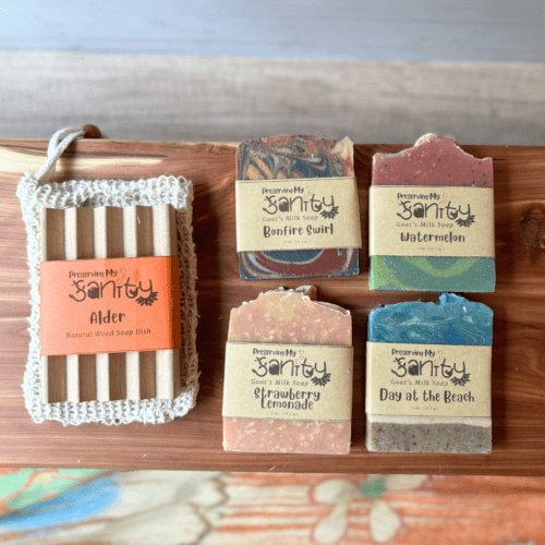 Flatlay photo sohwing the four half-size bars that are in a summer soap sampler, pictured in an arrangement with a sisal soap saver bag and alder natural wood soap dish. In this photo, soaps are shown with labels, so middle part of designs are hidden.