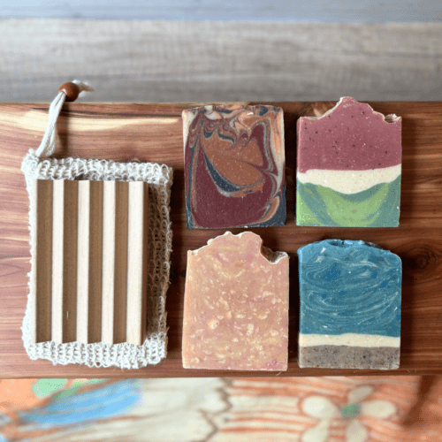 Flatlay photo sohwing the four half-size bars that are in a summer soap sampler, pictured in an arrangement with a sisal soap saver bag and alder natural wood soap dish. In this photo, the soaps are shown without labels so customers can see full design.