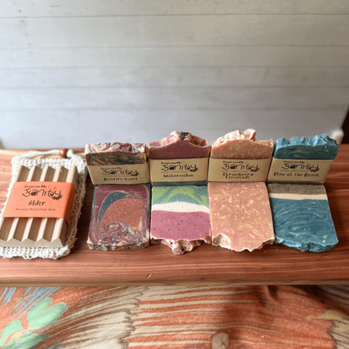 Alternate view of the soaps included in the summer soap sampler. This photo shows 8 half bars of soap - one of each kind in the sampler with label, and one without label. The sampler only includes 4 half size bars total, not all 8 as shown in this photo. Also included is a sisal soap saver bag and natural wood soap dish.