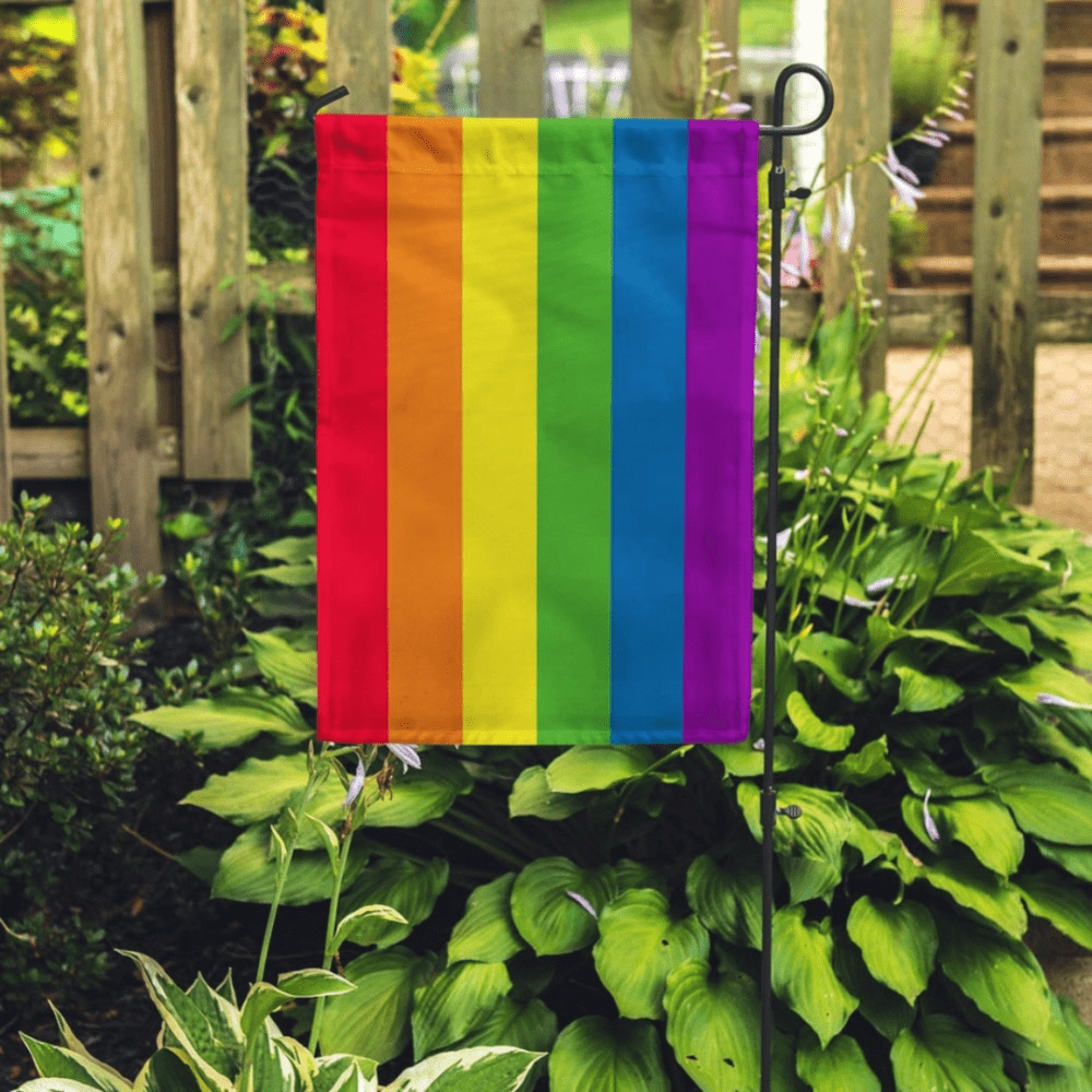 Display photo showcasing a rainbow pride garden flag on a stake in the garden, with pretty hostas and a wood fence in the background