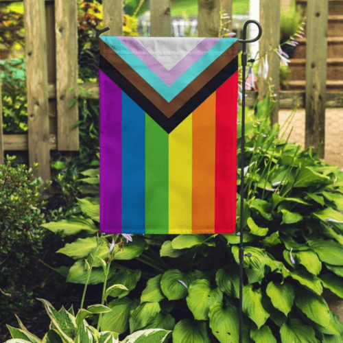 Progress pride garden flag displayed on a garden flag stake with pretty hostas and a wood fence in the background