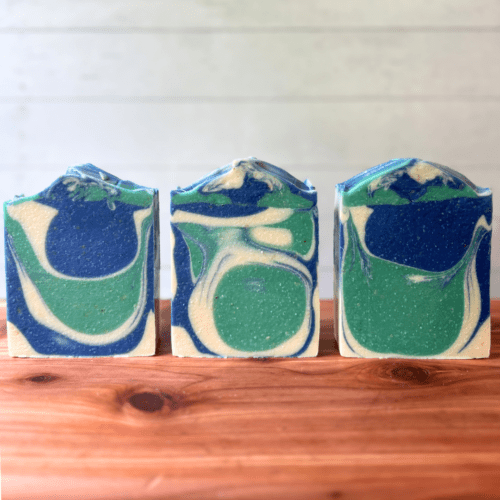 Closeup photo showing three bars of Cotton and Eucalyptus goat milk soap on a finished cedar board with a whitewashed wood background