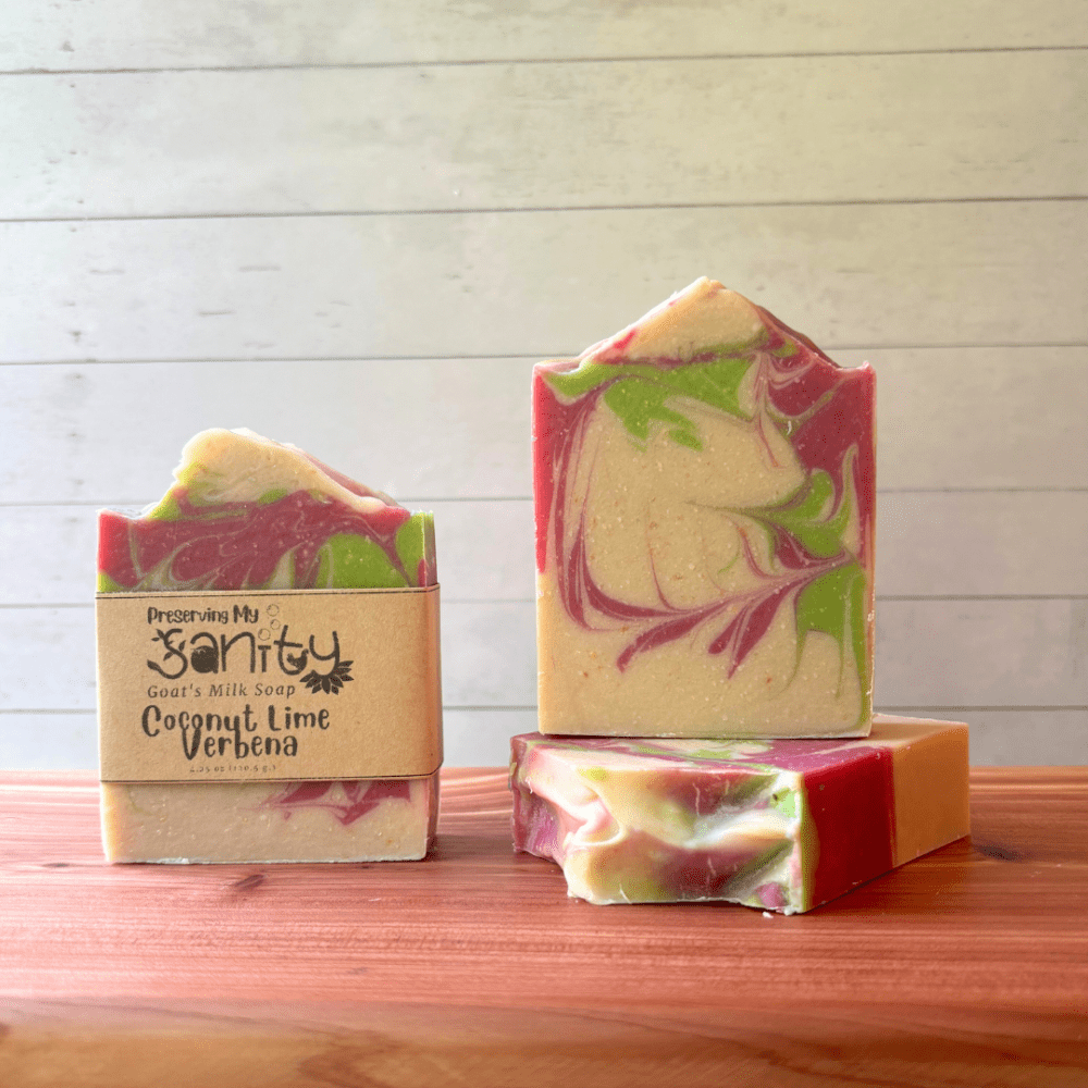 Three bars of Coconut Lime Verbena goat milk soap pictured to show the top, front, and label of this unique handcrafted soap. Sitting on a cedar board with a white wood background.