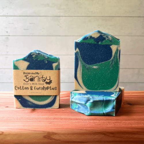 Three bars of Cotton & Eucalyptus goat milk soap pictured to show the top, front, and label of this unique handcrafted soap. Sitting on a cedar board with a white wood background.