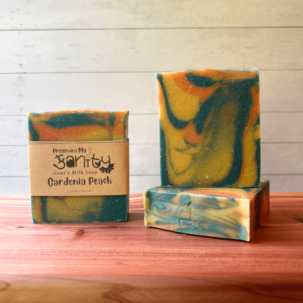 Three bars of Gardenia Peach goat milk soap pictured to show the top, front, and label of this unique handcrafted soap. Sitting on a cedar board with a white wood background.