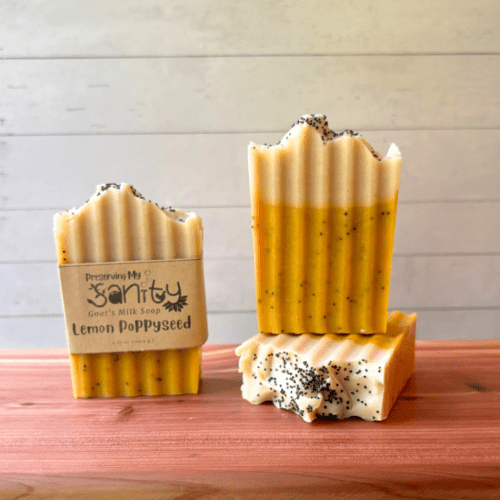 Three bars of Lemon Poppyseed goat milk soap pictured to show the top, front, and label of this unique handcrafted soap. Sitting on a cedar board with a white wood background.