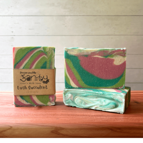 Three bars of Lush Succulent goat milk soap pictured to show the top, front, and label of this unique handcrafted soap. Sitting on a cedar board with a white wood background.