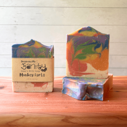 Three bars of Monkey Farts goat milk soap pictured to show the top, front, and label of this unique handcrafted soap. Sitting on a cedar board with a white wood background.