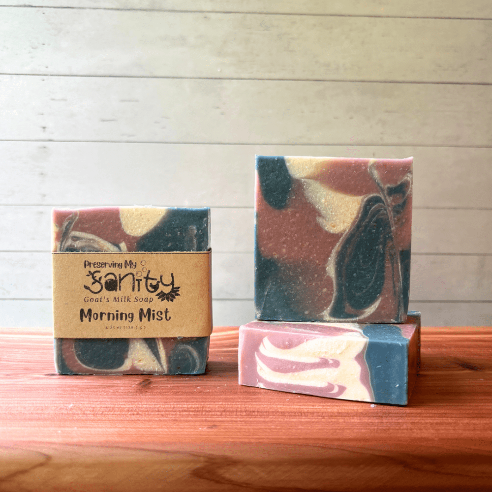 Three bars of Morning Mist goat milk soap pictured to show the top, front, and label of this unique handcrafted soap. Sitting on a cedar board with a white wood background.