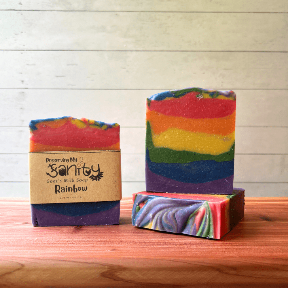 Three bars of Rainbow goat milk soap pictured to show the top, front, and label of this unique handcrafted soap. Sitting on a cedar board with a white wood background.