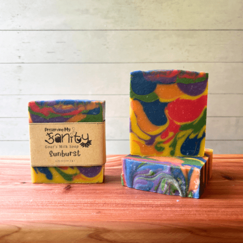 Three bars of Sunburst goat milk soap pictured to show the top, front, and label of this unique handcrafted soap. Sitting on a cedar board with a white wood background.