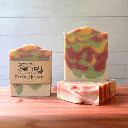 Three bars of Tropical Breeze goat milk soap pictured to show the top, front, and label of this unique handcrafted soap. Sitting on a cedar board with a white wood background.