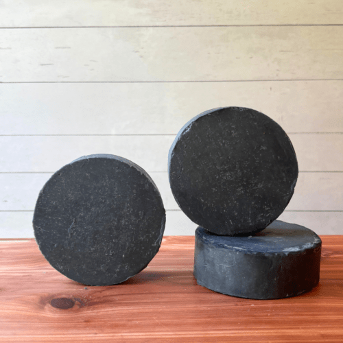 Display photo showcasing three round bars of charcoal and tea tree goat milk soap, handcrafted in small batches with local Minnesota goat milk.