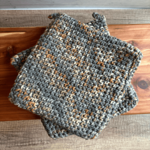 Alternate flatlay view of two crocheted potholders with gray as the main color stitched with a natural stone variegated yarn.