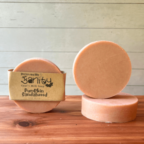 Display photo showcasing three round bars of Pumpkin Sandalwood goat milk soap. Two bars are plain without a label and show different angles of the soap, while the third bar features a company label.