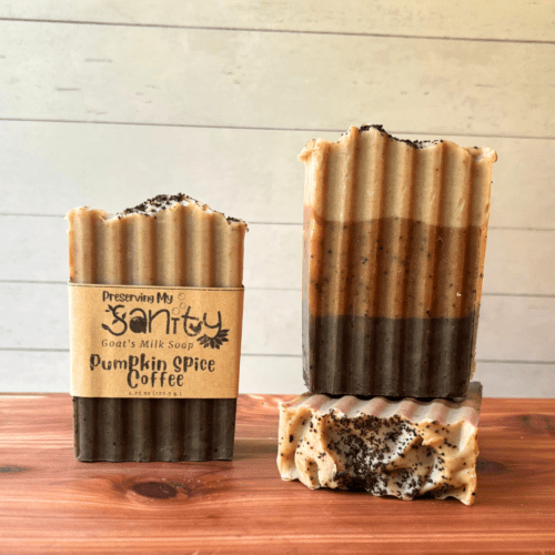 Front angle view of three bars of Pumpkin Spice Coffee goat milk soap, one showing a label and the other two showing different sides of the soap. The soap is a three-shade layered brown soap with coffee grounds on top.