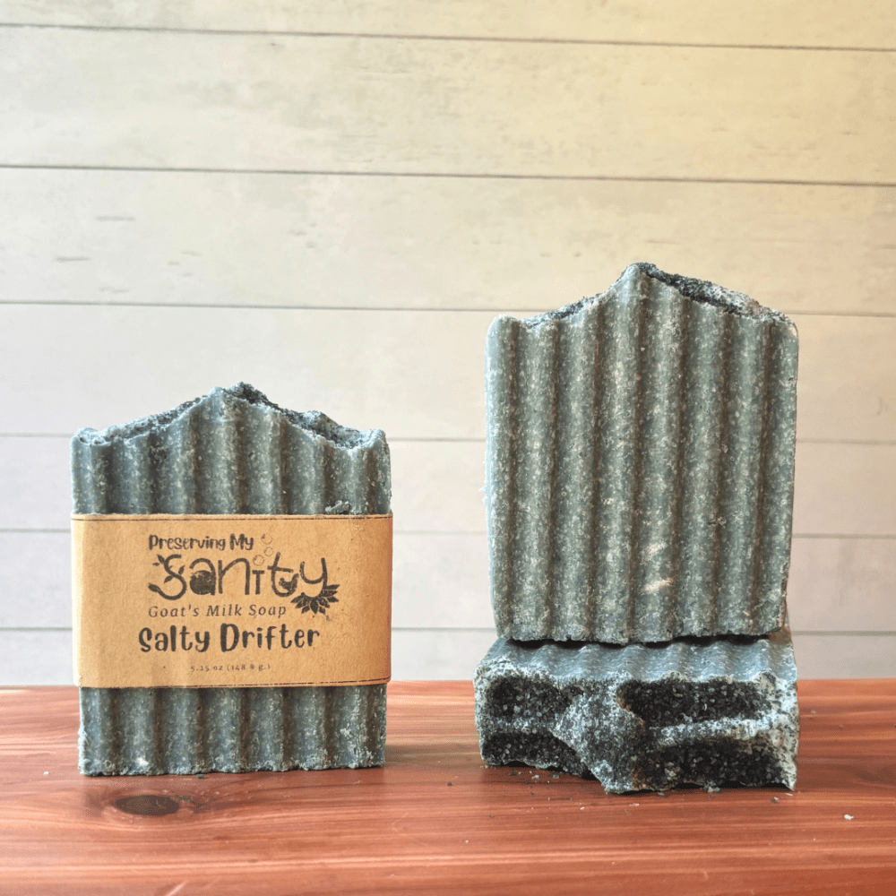 Front view of three bars of Salty Drifter sea salt soap, made with a mix of Hawaiian black soap and regular sea salt. The bars are a dark gray color with a hint of white swirls. One bar shows a soap label, the other two are displayed to show different angles of the soap. This handcrafted goat milk soap is handcrafted in small batches in Minnesota with local goat milk.