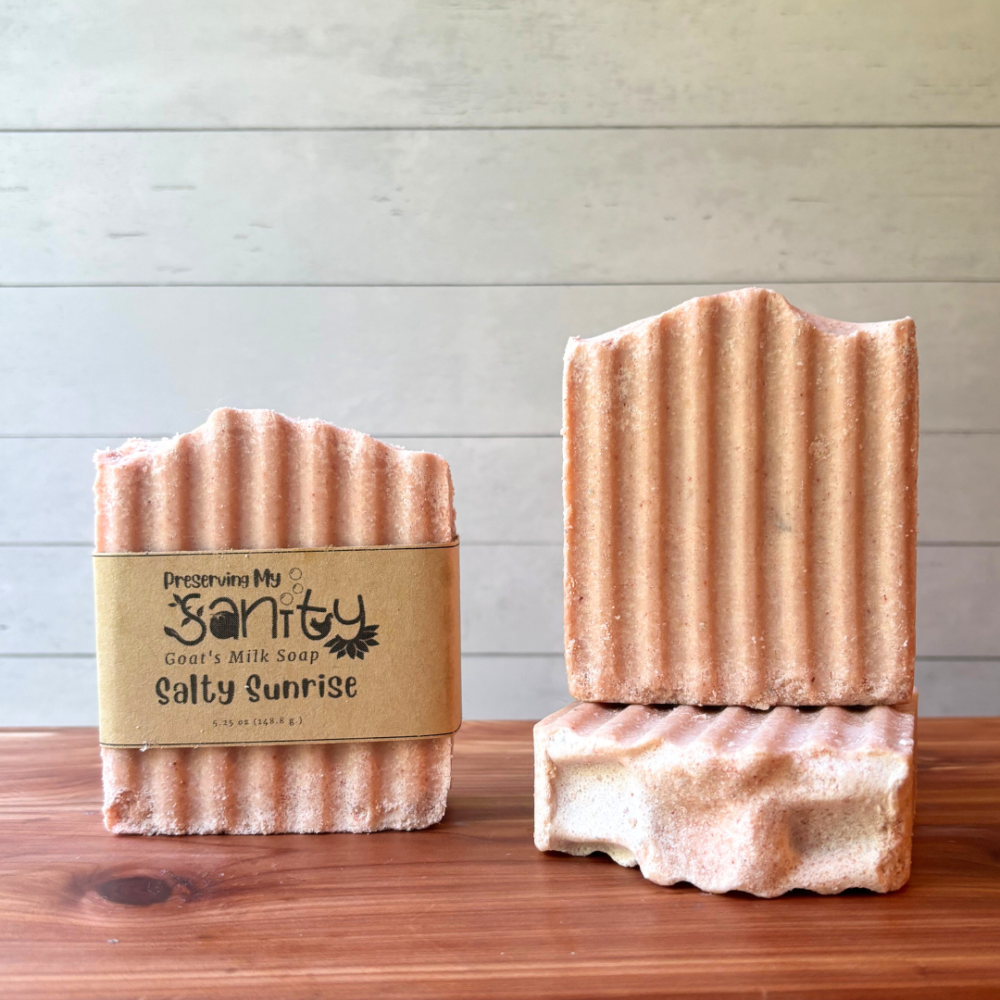 Front view of three bars of Salty Sailor Pink Himalayan sea salt soap. One bar shows a soap label, the other two are displayed to show different angles of the soap. The bars are a coral pink color with a hint of white swirls and scented in a floral grapefruit scent. This handcrafted goat milk soap is handcrafted in small batches in Minnesota with local goat milk.