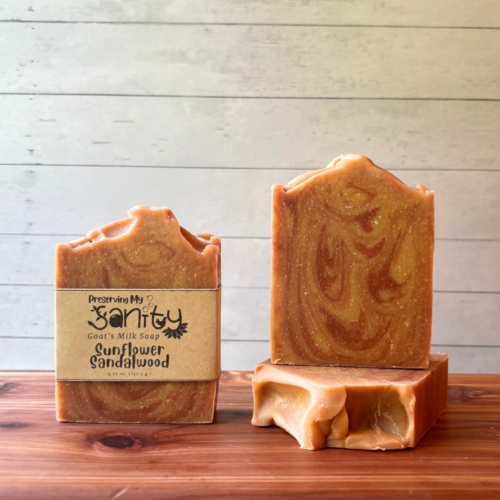 This photo showcases three bars of Sunflower Sandalwood goat milk soap, handcrafted in small batches with local Minnesota goat milk. There are three bars of soap in the photo, one showing the label and the two positioned to show alternate angles of these dark golden brown and beige swirled soap bars.