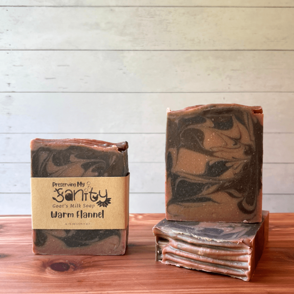 Display photo showcasing three bars of Warm Flannel soap as part of the fall collection. Soap is handcrafted in small batches with local Minnesota goat milk. These bars are swirled with light brown, dark brown, and dark gray and the top was textured with tines of a fork. The photo shows one bar with a label and the other two without a label to show different sides of these pretty artisan soap bars.