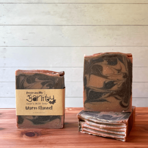 Display photo showcasing three bars of Warm Flannel soap as part of the fall collection. Soap is handcrafted in small batches with local Minnesota goat milk. These bars are swirled with light brown, dark brown, and dark gray and the top was textured with tines of a fork. The photo shows one bar with a label and the other two without a label to show different sides of these pretty artisan soap bars.