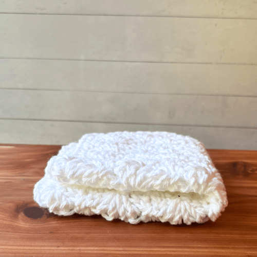 Alternate side view of a pretty white crocheted washcloth folded in quarters sitting on a cedar bath board.