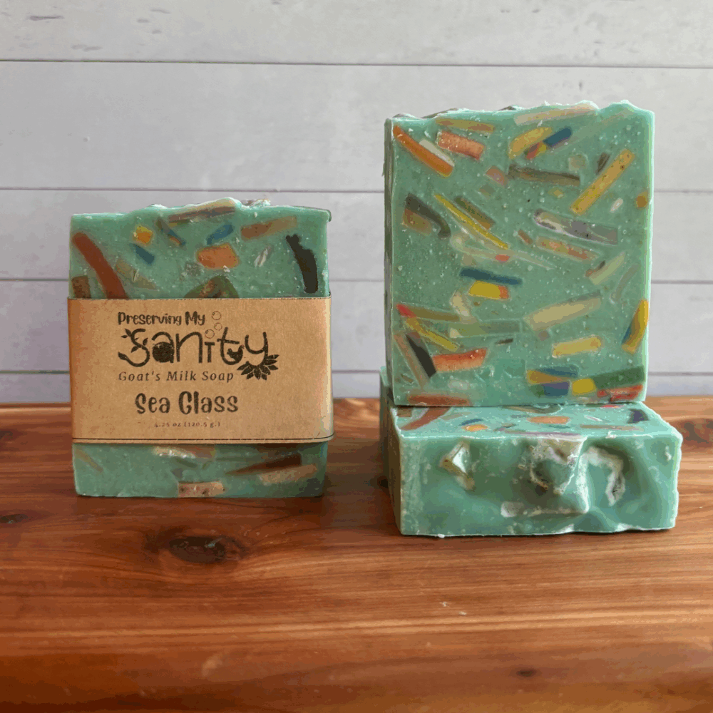 Three bars of Sea Glass confetti soap. The soap is a light aqua blue color with slices of various colors of goat milk soap embedded into it to look like sea glass. The scent is a clean beachy fragrance. The soaps are pictured on a cedar bath board with a white paneled background.