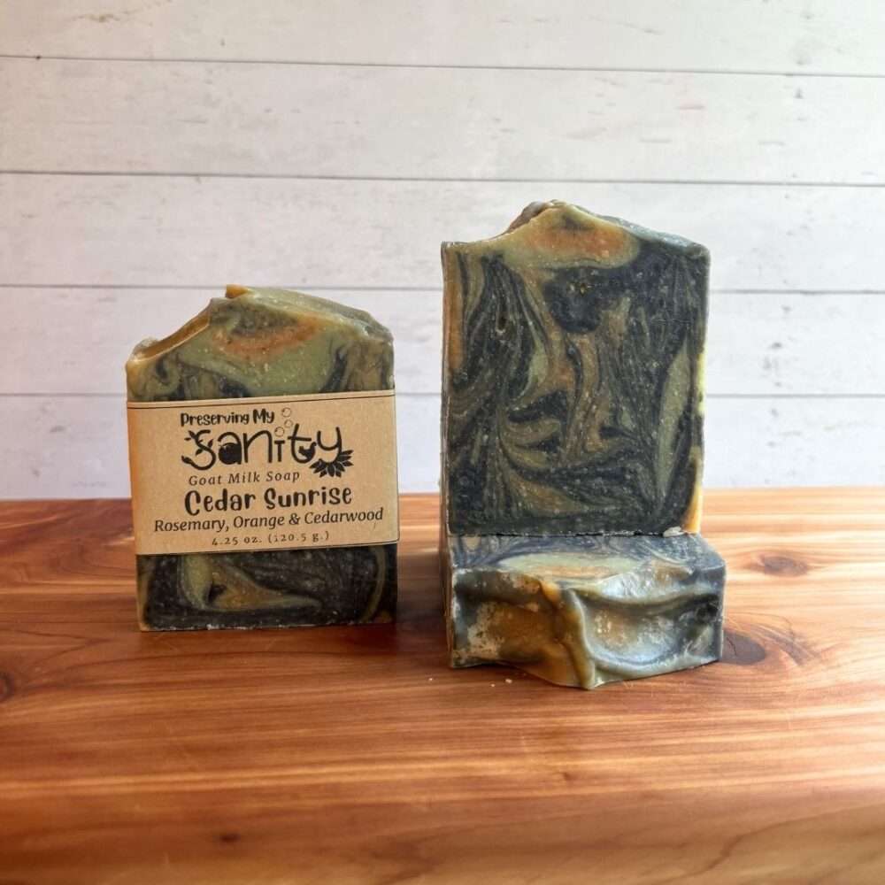 Three bars of Cedar Sunrise Goat Milk Soap are displayed on a warm cedar wood bath board against a white shiplap background. One bar stands upright with the signature kraft paper label reading "Cedar Sunrise." A second unwrapped bar stands upright beside it, showcasing dramatic marbled swirls of deep charcoal, mossy green, warm olive, and golden amber. A third unwrapped bar lies flat in front, revealing the textured top with the same flowing artisan swirl pattern.