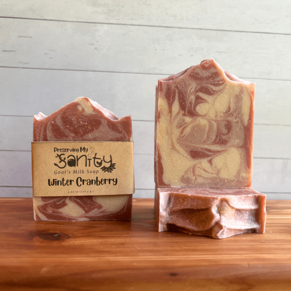 Three bars of Winter Cranberry goat milk soap sitting on a cedar bath board with a white paneled background. One bar is shown without a label to display the swirl design, while one other bar shows the textured top and the third is displayed with a label on it.
