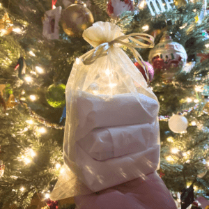 Holiday promo photo highlighting a holiday goat milk soap grab bag in a gold organza gift bag, being held up in front of a classy decorated Christmas tree with pretty ornaments, beaded gardland, and white lights.