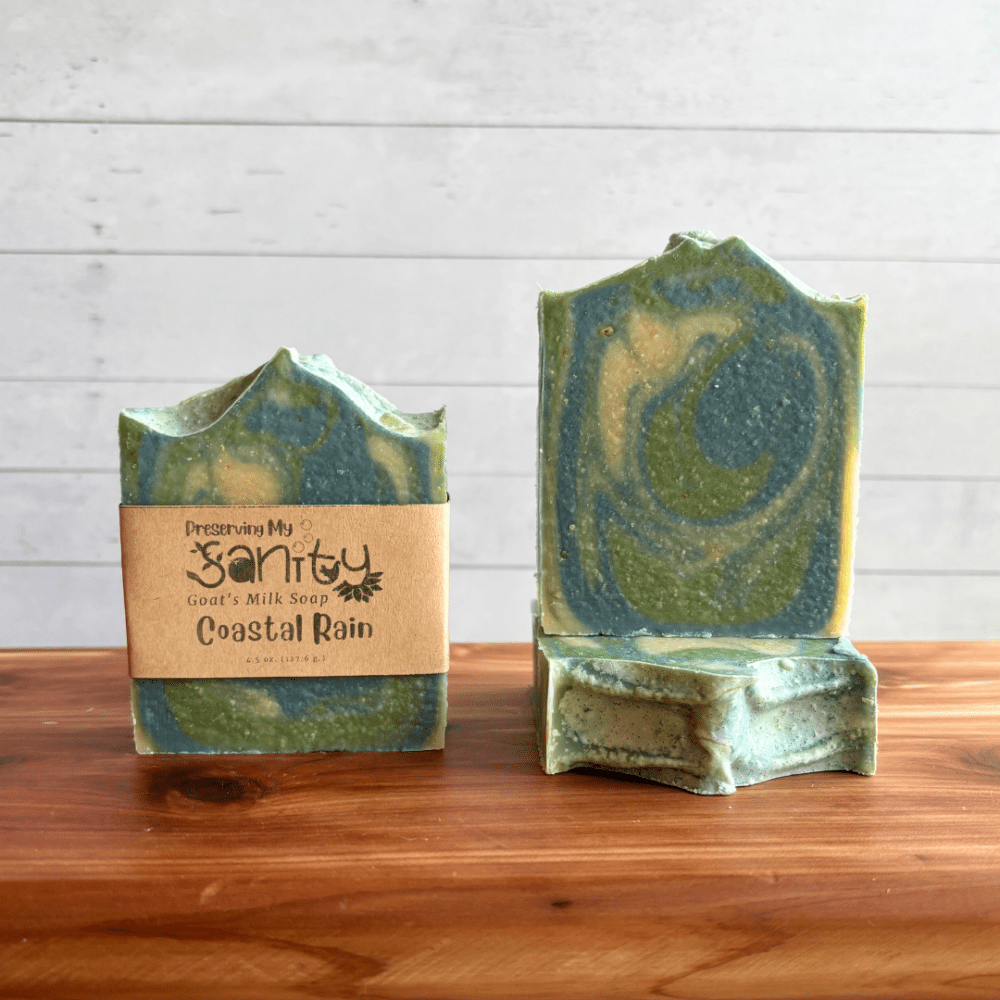 Display photo showcasing three bars of Coastal Rain goat milk soap, one with a label and one without a label to showcase the soap design. A third bar of soap is underneath that showcasing the top of the bar of soap. The bars of soap are sitting on a cedar bath board with a white shiplap background behind it.