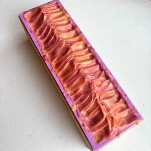 Process photo showing one loaf of Coconut Paradise vegan coconut milk soap. The top of the soap has been textured with a spoon and you can see the hot pink, orange and yellow colors that are swirled within the soap.