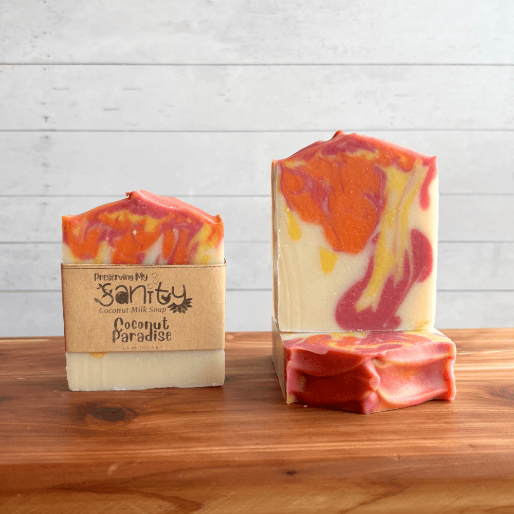 Display photo showcasing three bars of Coconut Paradise vegan coconut milk soap, one with a label and one without a label to showcase the soap design. A third bar of soap is underneath that showcasing the top of the bar of soap. The bars of soap are sitting on a cedar bath board with a white shiplap background behind it.