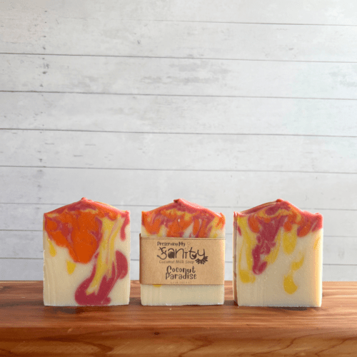 Display photo showcasing three bars of Coconut Paradise vegan coconut milk soap, one with a label and two without labels to showcase soap design. The bars of soap are sitting on a cedar bath board with a white shiplap background behind it.