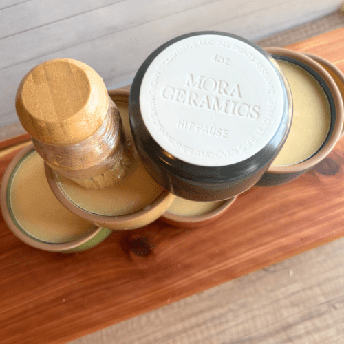 Photo of several ceramic ramekins of dish soap pictured with a bamboo dish brush. The top ramekin is pictured upside down to show the pottery brand, Mora Ceramics.