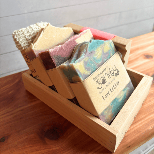 Display photo showcasing three bars of goat milk soap, a natural wood soap dish, and a soap saver bag situated in a wood gift tray. Set is sitting on a cedar bath board.