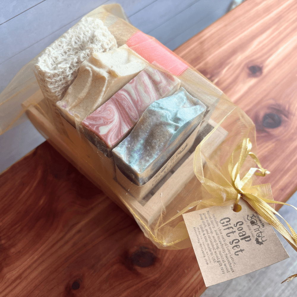 Display photo showcasing three bars of goat milk soap, a natural wood soap dish, and a soap saver bag situated in a wood gift tray and packaged in a gold organza gift bag with a tag on the outside. Gift set is sitting on a cedar bath board.