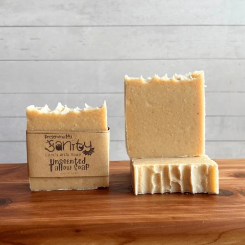 Display photo showcasing three bars of plain unscented Beef Tallow Soap with Goat Milk, one with a label and one without a label to showcase the soap design. A third bar of soap is underneath that showcasing the top of the bar of soap. The bars of soap are sitting on a cedar bath board with a white shiplap background behind it.