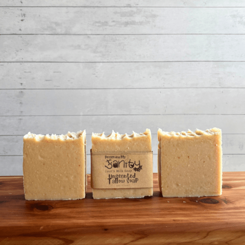 DIsplay photo showcasing three bars of plain unscented Tallow Goat Milk Soap, one with a label and two without labels to showcase soap design. The bars of soap are sitting on a cedar bath board with a white shiplap background behind it.