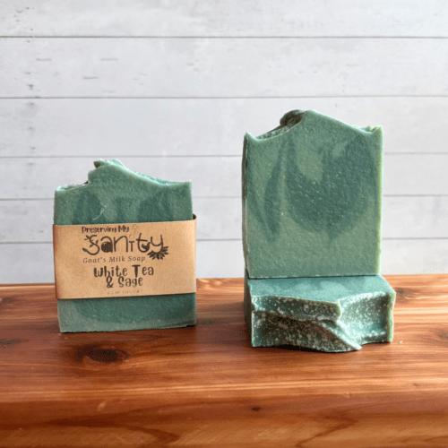 DIsplay photo showcasing three bars of White Tea and Sage goat milk soap, one with a label and one without a label to showcase the soap design. A third bar of soap is underneath that showcasing the top of the bar of soap. The bars of soap are sitting on a cedar bath board with a white shiplap background behind it.