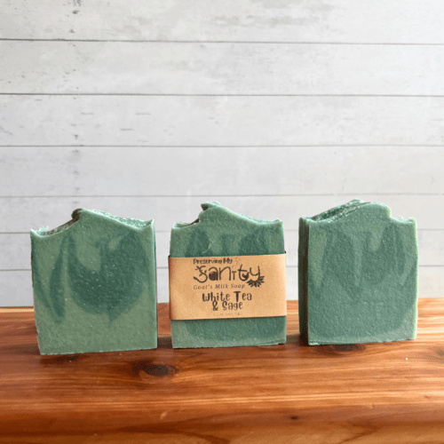 DIsplay photo showcasing three bars of White Tea and Sage goat milk soap, one with a label and two without labels to showcase soap design. The bars of soap are sitting on a cedar bath board with a white shiplap background behind it.