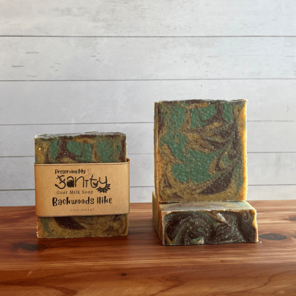 Alternate view display photo showcasing three bars of Backwoods Hike goat milk soap sitting on a cedar board with a white shiplap background. This soap has a yellow base and is swirled with aqua blue and brown. It also contains walnut shell powder for exfoliation, so you can see some brown specks in the soap.