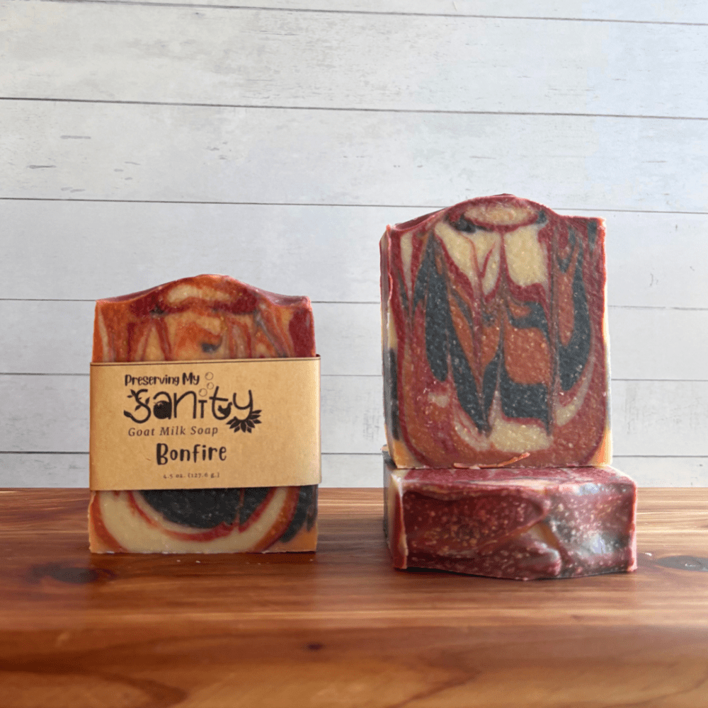 Alternate view display photo showcasing three bars of Bonfire goat milk soap sitting on a cedar board with a white shiplap background. This soap is my bestseller and features a unique swirled pattern of black, red, orange, and white.